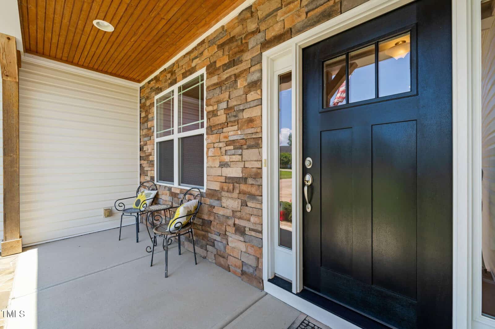 A front porch with a black door, stone and siding exterior walls, two metal chairs with yellow cushions, and a concrete floor. A window is next to the door, and the ceiling is wood-paneled.