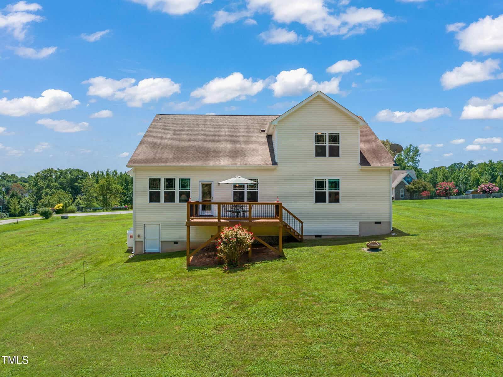 A two-story white house with a wooden deck sits on a large, well-maintained grassy lawn under a blue sky with scattered clouds. A small bush is at the base of the deck, and a fire pit is in the yard.
