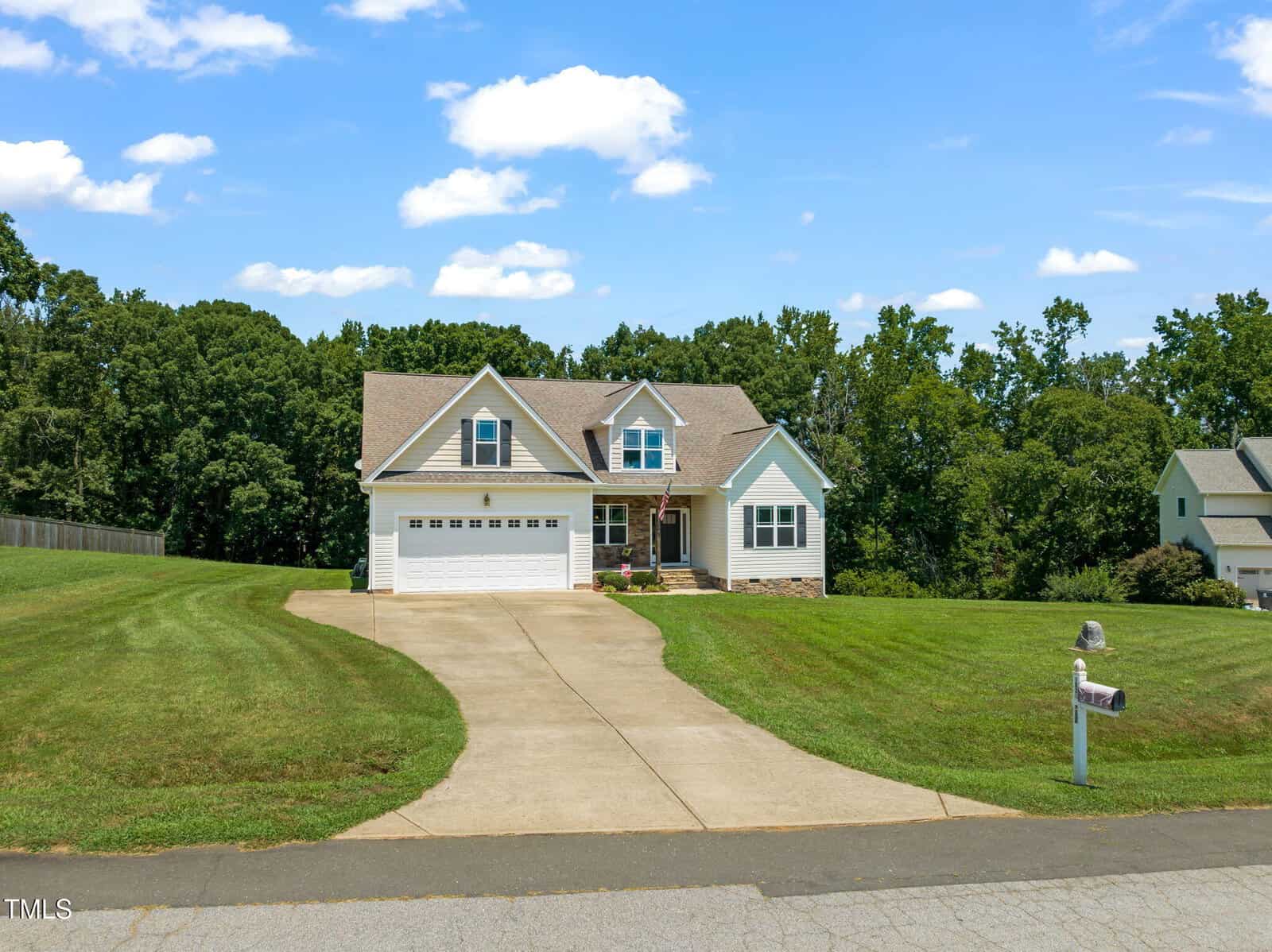 Located at 1034 Snapdragon Drive in Wake Forest, this two-story suburban house features an attached three-car garage, beige siding, a large sloped driveway, and a well-maintained green lawn bordered by trees under a blue sky.