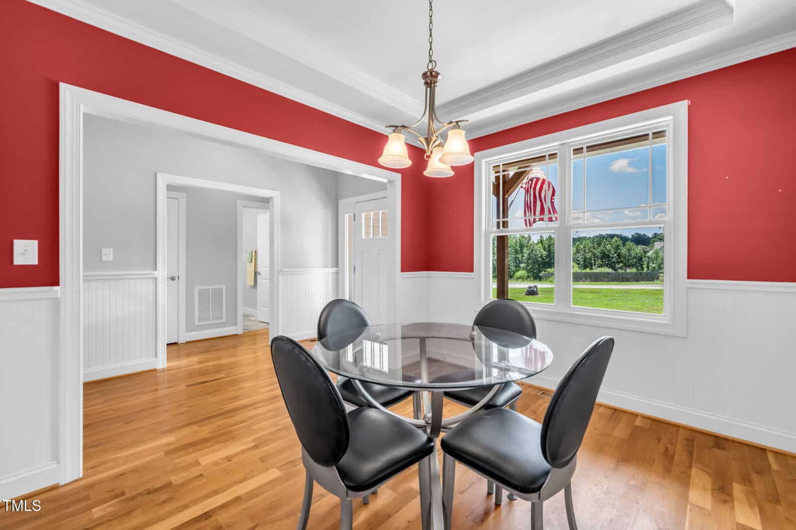 A dining area with red and white walls, a glass-top round table, and four black chairs. A window shows a green lawn and American flag outside. Hardwood floors and a ceiling light fixture are visible.
