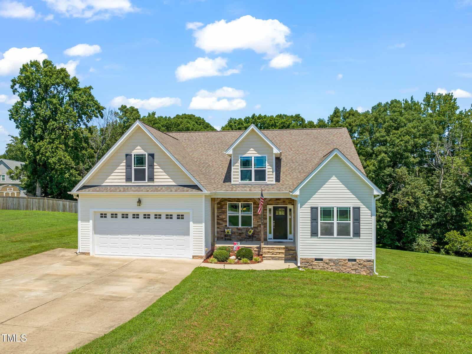 A two-story suburban house with beige siding, stone accents, a gable roof, a front porch, double garage, and a well-maintained lawn, set against a backdrop of trees under a blue sky with scattered clouds.