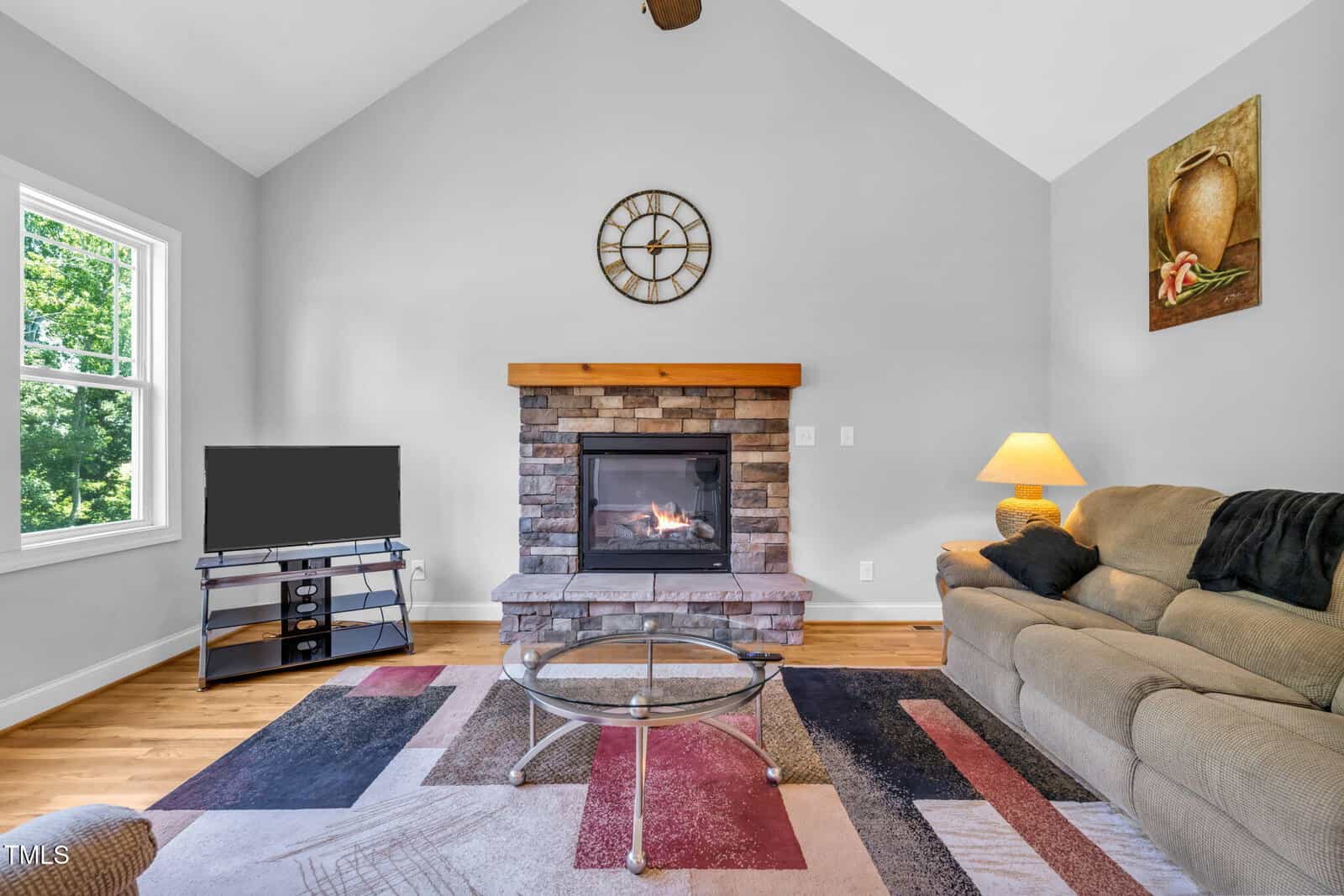 A living room with a vaulted ceiling features a stone fireplace, a wall clock above it, a TV on a stand, a beige sofa with a lamp, a glass coffee table, and a multicolored rug on a wooden floor.