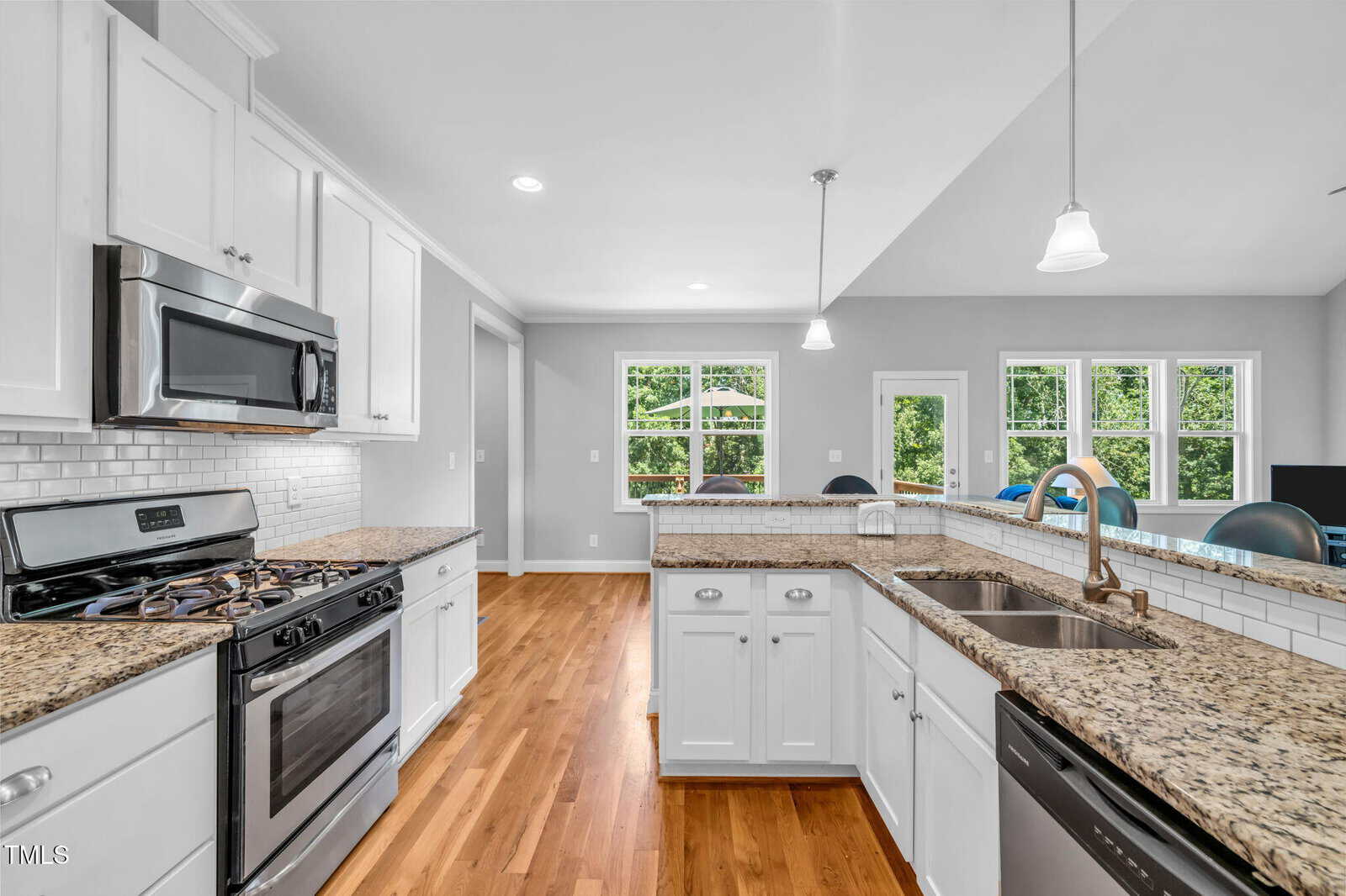 Bright kitchen with white cabinets, granite countertops, stainless steel appliances, a gas stove, wood floors, and large windows letting in natural light. A breakfast bar separates the kitchen from the living area.