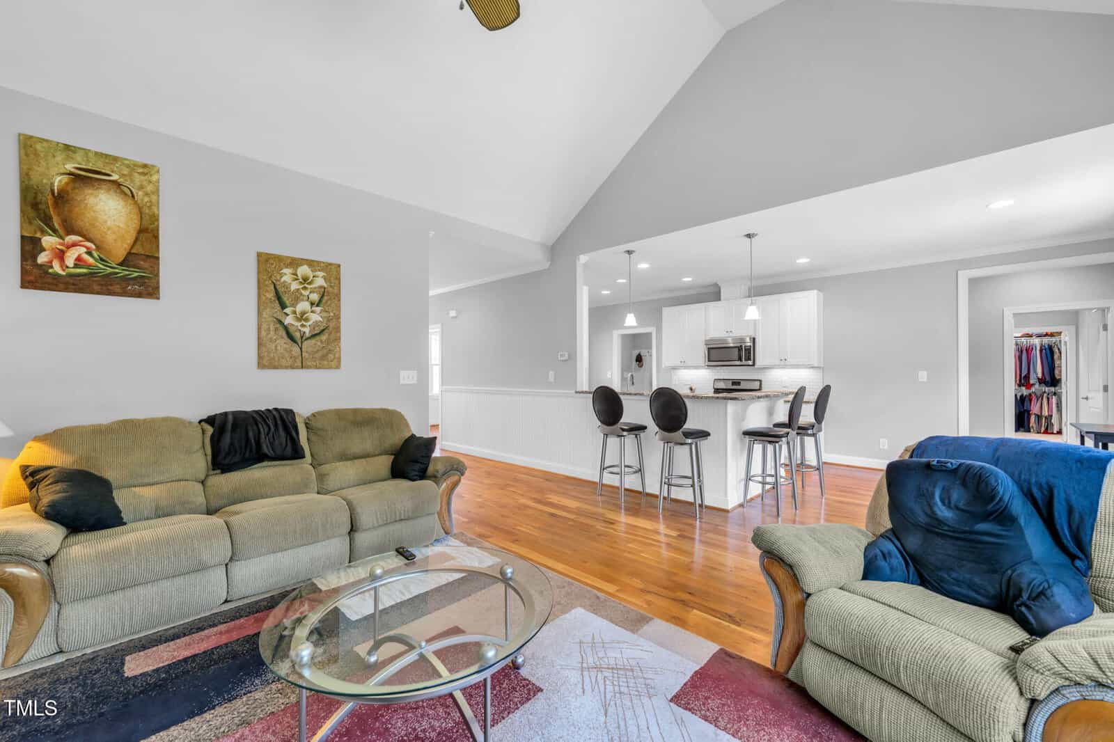 A spacious living room with a glass coffee table, two sofas, hardwood floors, and a view into a modern white kitchen with barstools. Two framed pictures hang on a gray wall above the couch.
