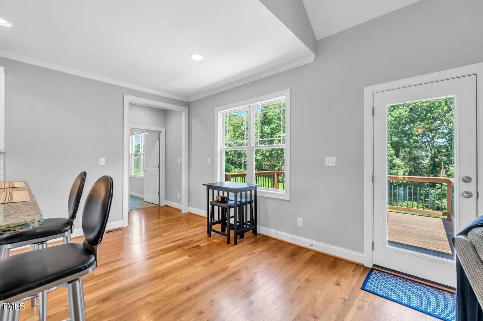 Bright dining area with wooden floors, a small black table and two chairs near a window, and a glass door leading to a deck with a view of green trees outside. Kitchen stools are partially visible on the left.