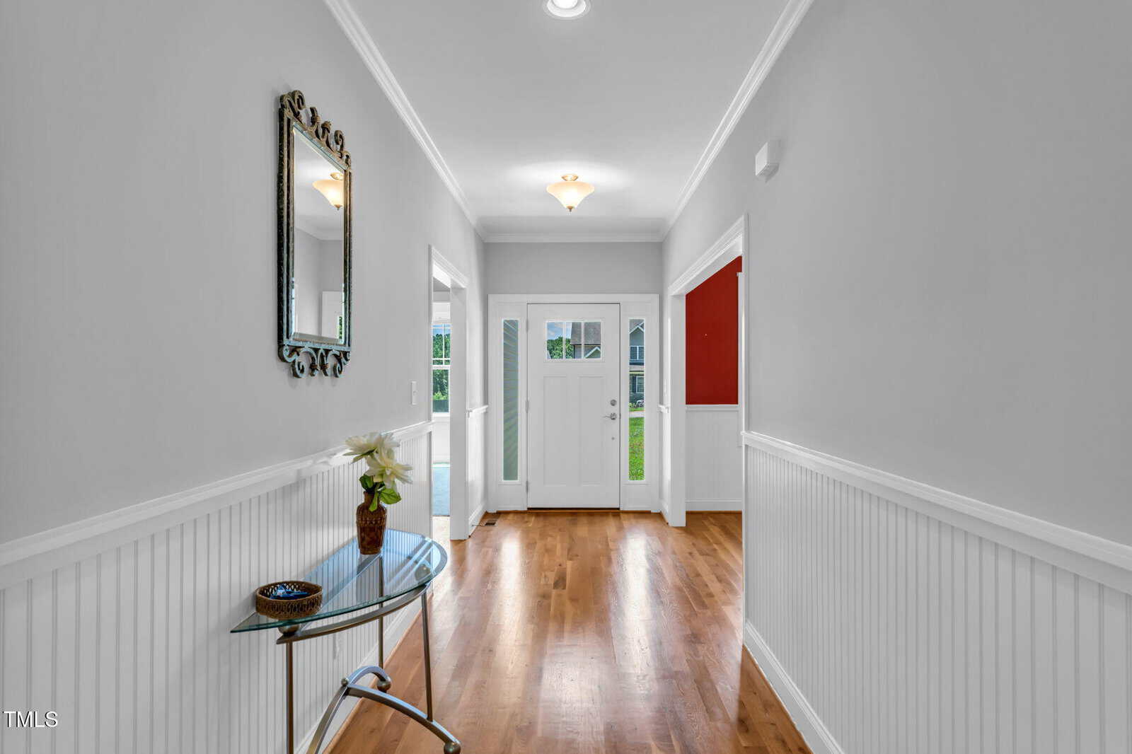 A hallway with wood flooring, gray walls, and white wainscoting. A glass table with a plant and a mirror are on the left wall. Double front doors with glass panels are at the end of the hall.