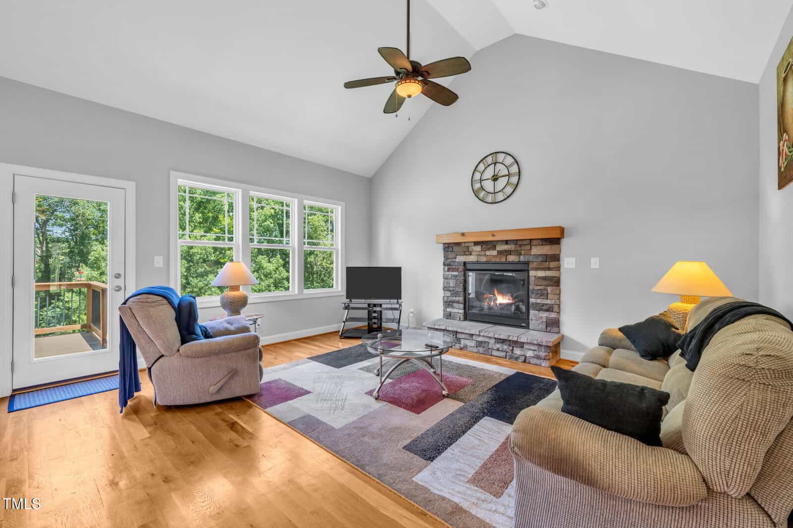 A living room with hardwood floors, a fireplace with a stone surround, two sofas, a glass coffee table, a TV, large windows, a ceiling fan, and a door leading to an outdoor deck.