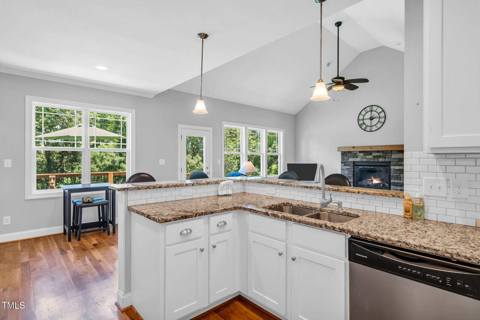 Bright kitchen with white cabinets, granite countertops, stainless appliances, and a double sink overlooks a living room with a fireplace and a dining area with large windows showcasing a green outdoor view.