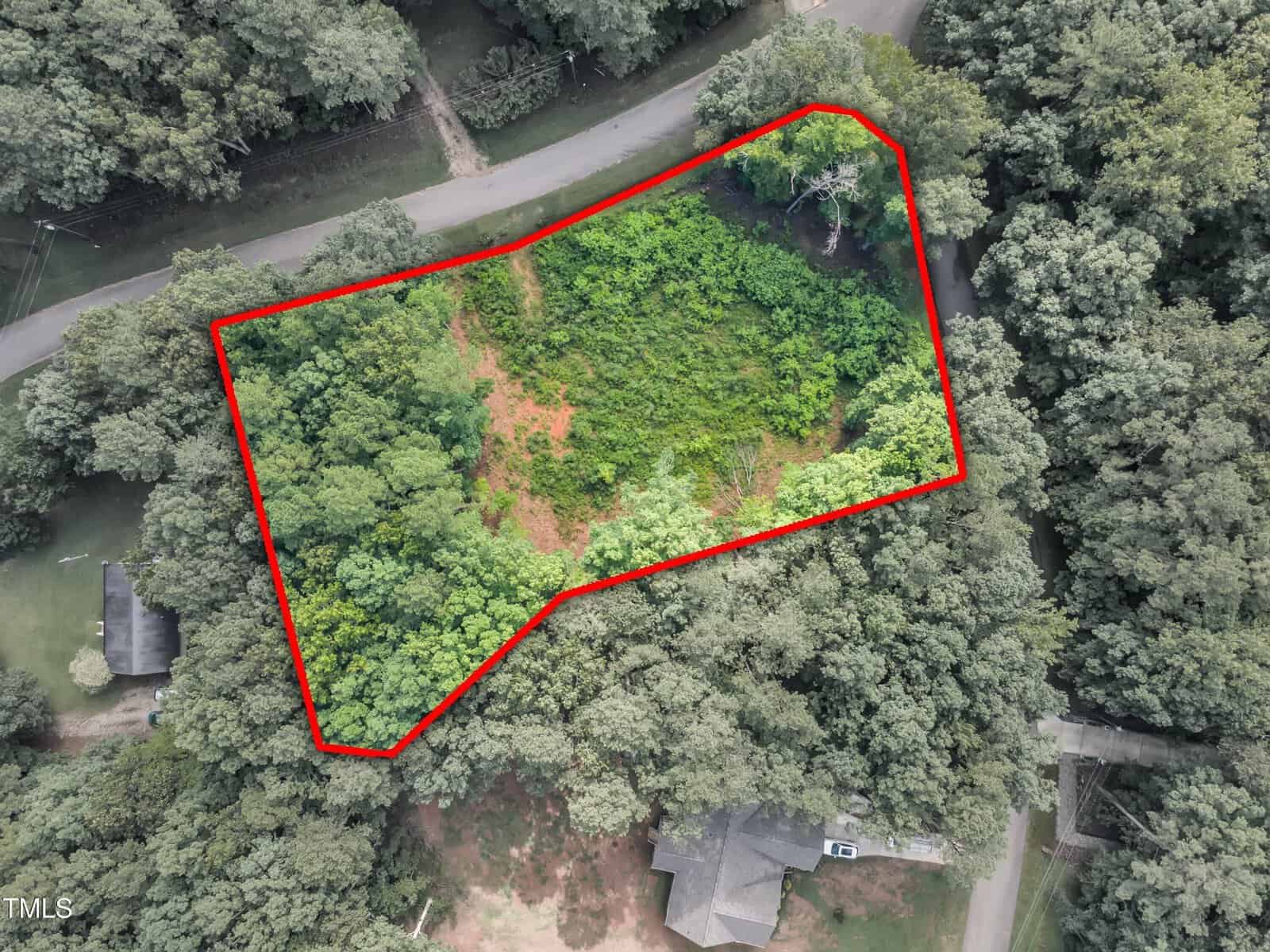 Aerial view of a tree-covered plot of land outlined in red, bordered by roads on two sides, with houses and dense forest surrounding the area.