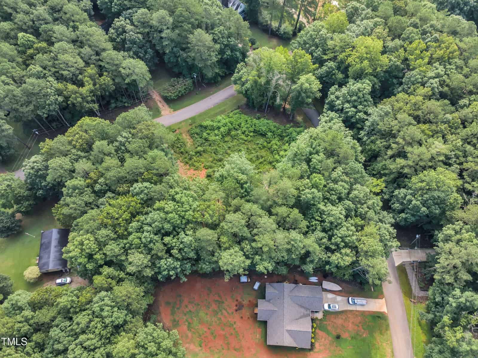 Aerial view of a house surrounded by trees with a driveway, nearby road, and another smaller house visible. The area has dense greenery and appears residential and forested.