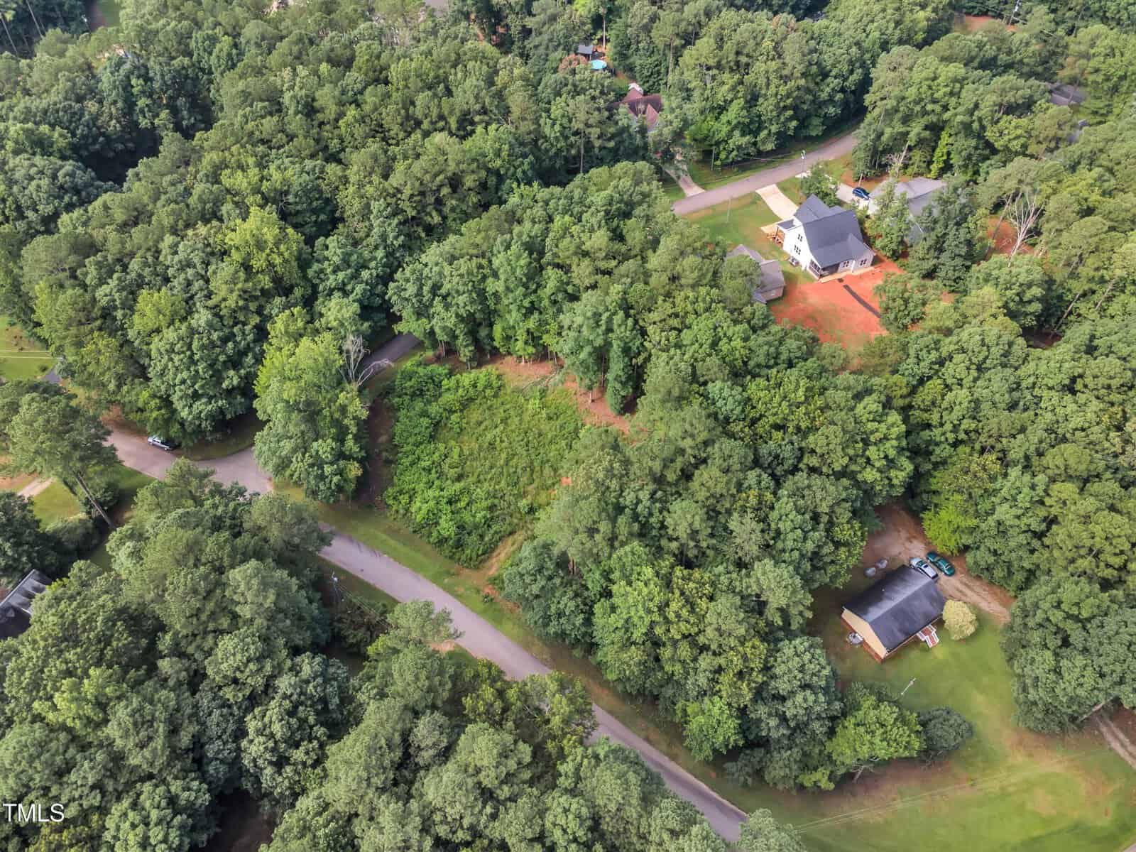 Aerial view of a wooded residential area with a few houses, curved roads, and dense green trees surrounding cleared patches of land.