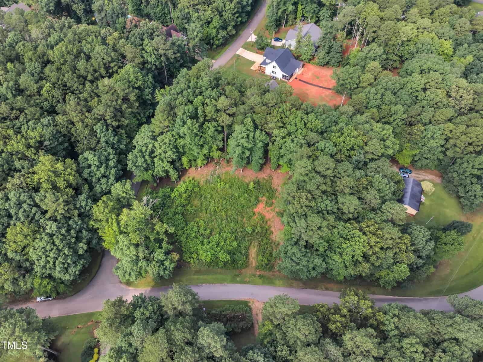 Aerial view of a wooded residential area with roads curving around a large, partially cleared grassy lot surrounded by dense trees. Nearby houses are visible among the trees.