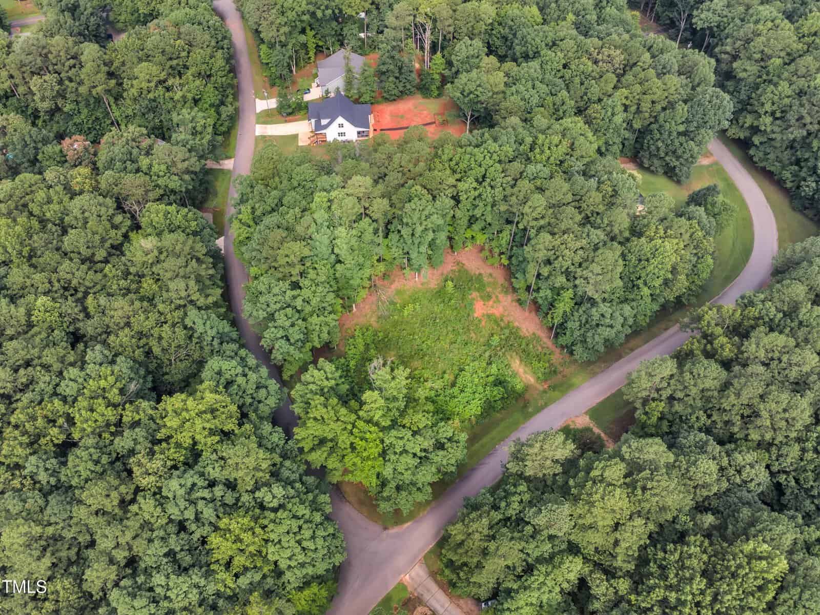 Aerial view of a wooded neighborhood with a cleared lot at the center bordered by curved roads. A white house with a driveway is visible near the top left, surrounded by dense green trees.
