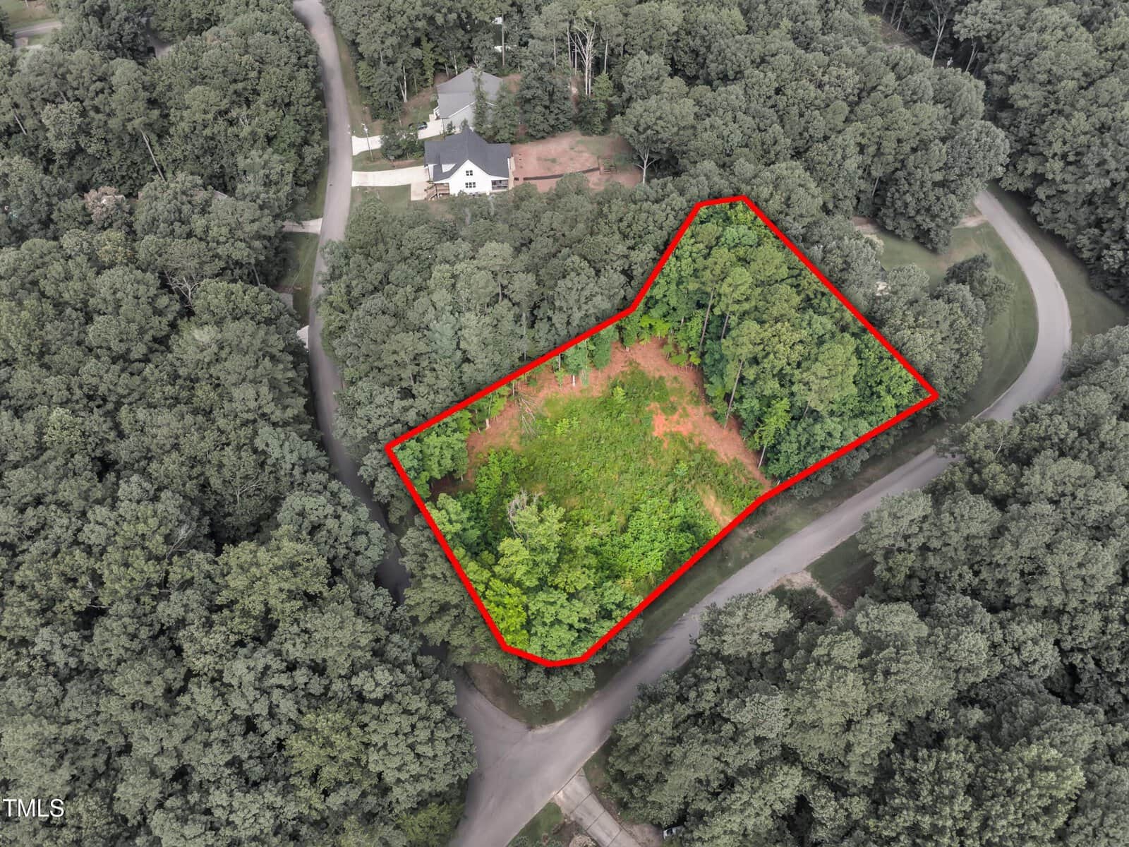 Aerial view of a wooded residential lot outlined in red, located at the intersection of two roads and surrounded by trees, with a nearby house visible at the top of the image.
