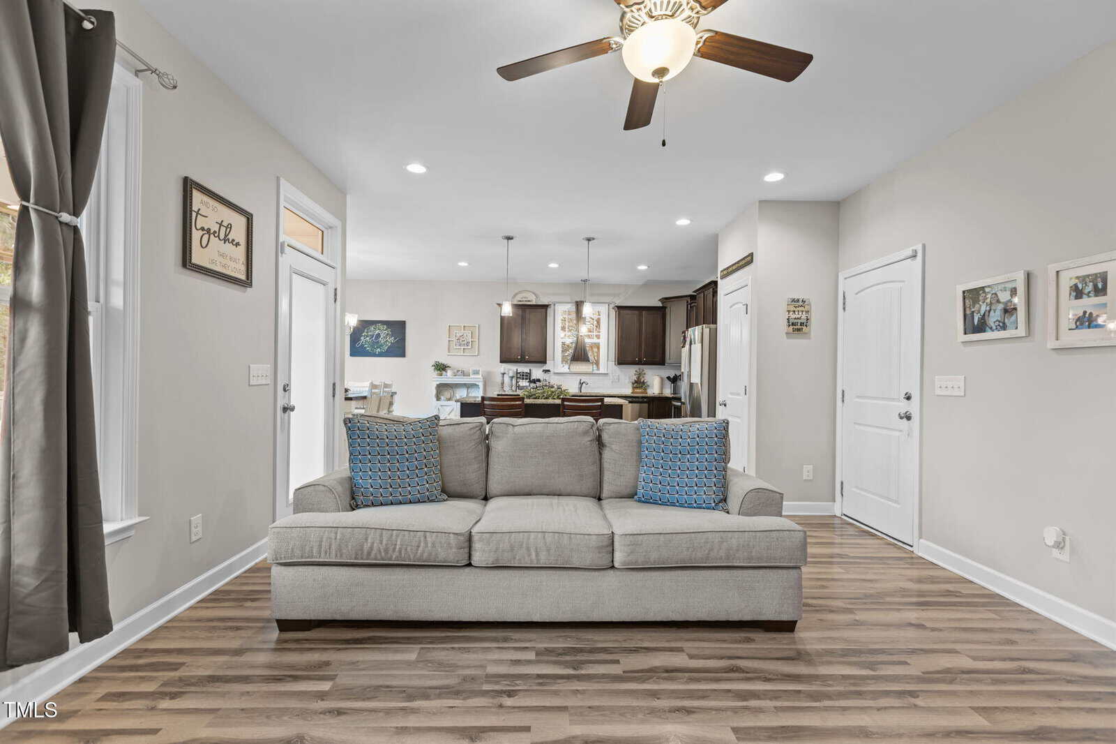 A living room with a light gray sofa featuring two blue patterned pillows, wood flooring, neutral walls, and a ceiling fan. The kitchen with dark cabinets and a dining area are visible in the background.