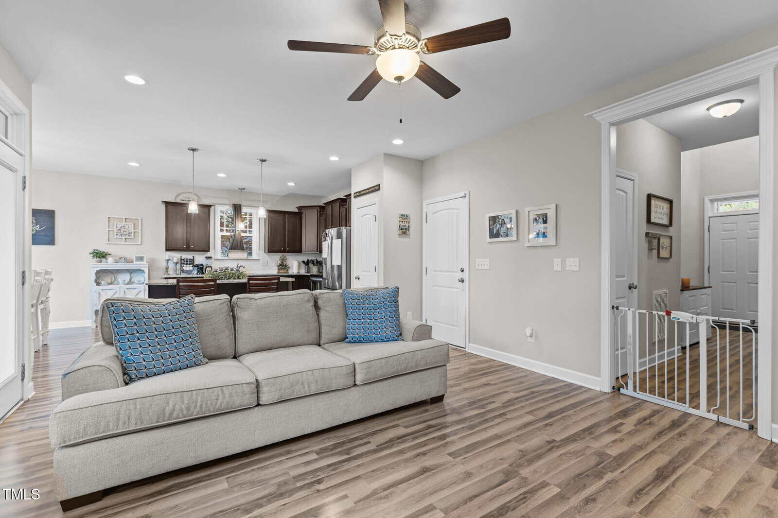 A spacious, open-concept living area with wood flooring, a gray sofa with blue patterned pillows, a ceiling fan, and a view of the kitchen with dark cabinets and pendant lights in the background.