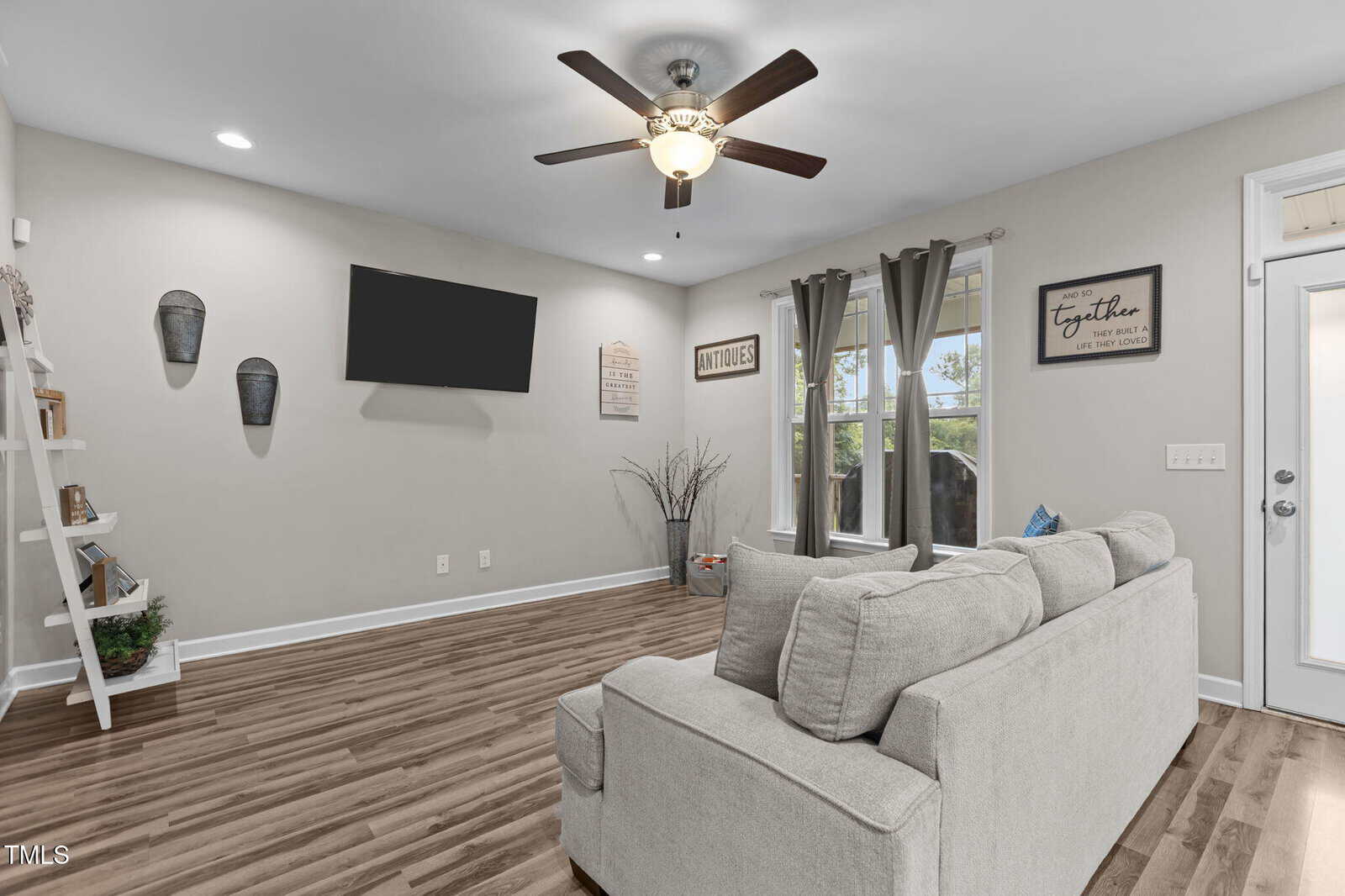 A living room with light gray walls, wood flooring, a ceiling fan with lights, a wall-mounted TV, and a gray sofa. Wall art, shelves, and two windows with gray curtains are visible.