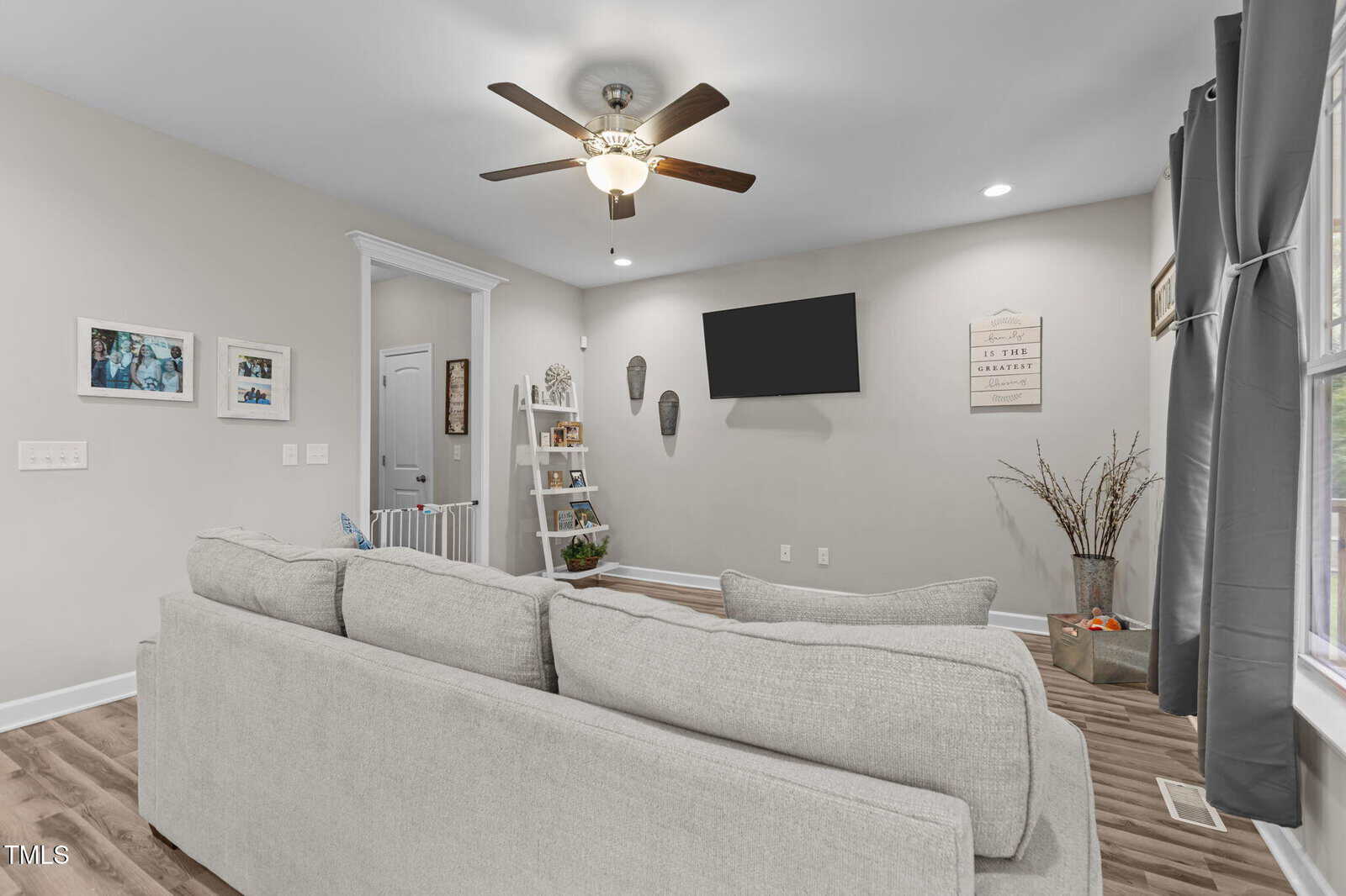 A living room with a gray sofa facing a wall-mounted TV. The room has wood flooring, a ceiling fan with lights, a small ladder shelf, framed photos on the wall, and large windows with gray curtains.