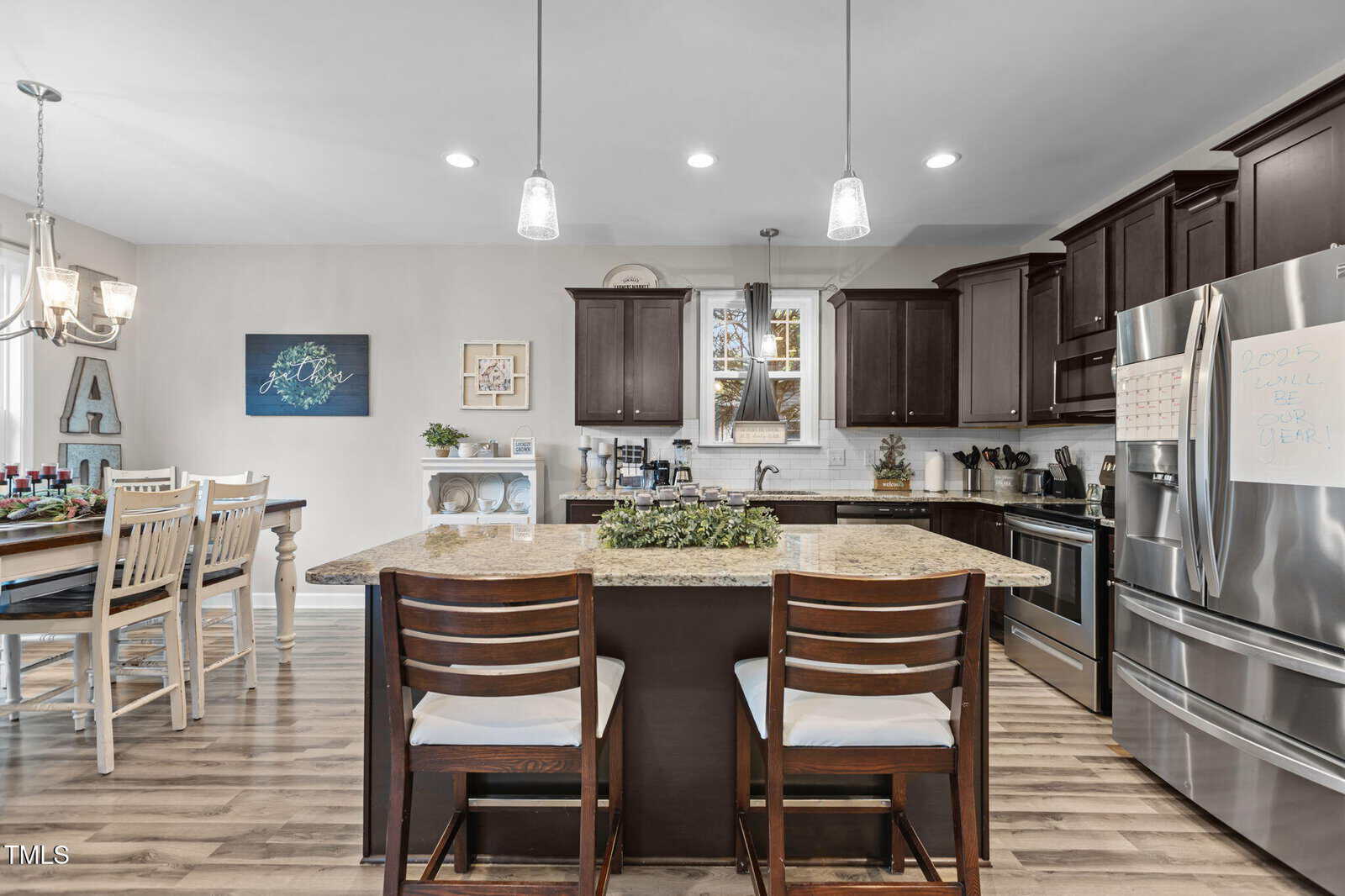 A modern kitchen with dark wood cabinets, granite countertops, stainless steel appliances, a central island with two chairs, pendant lighting, and a dining table set for four in the adjacent dining area.