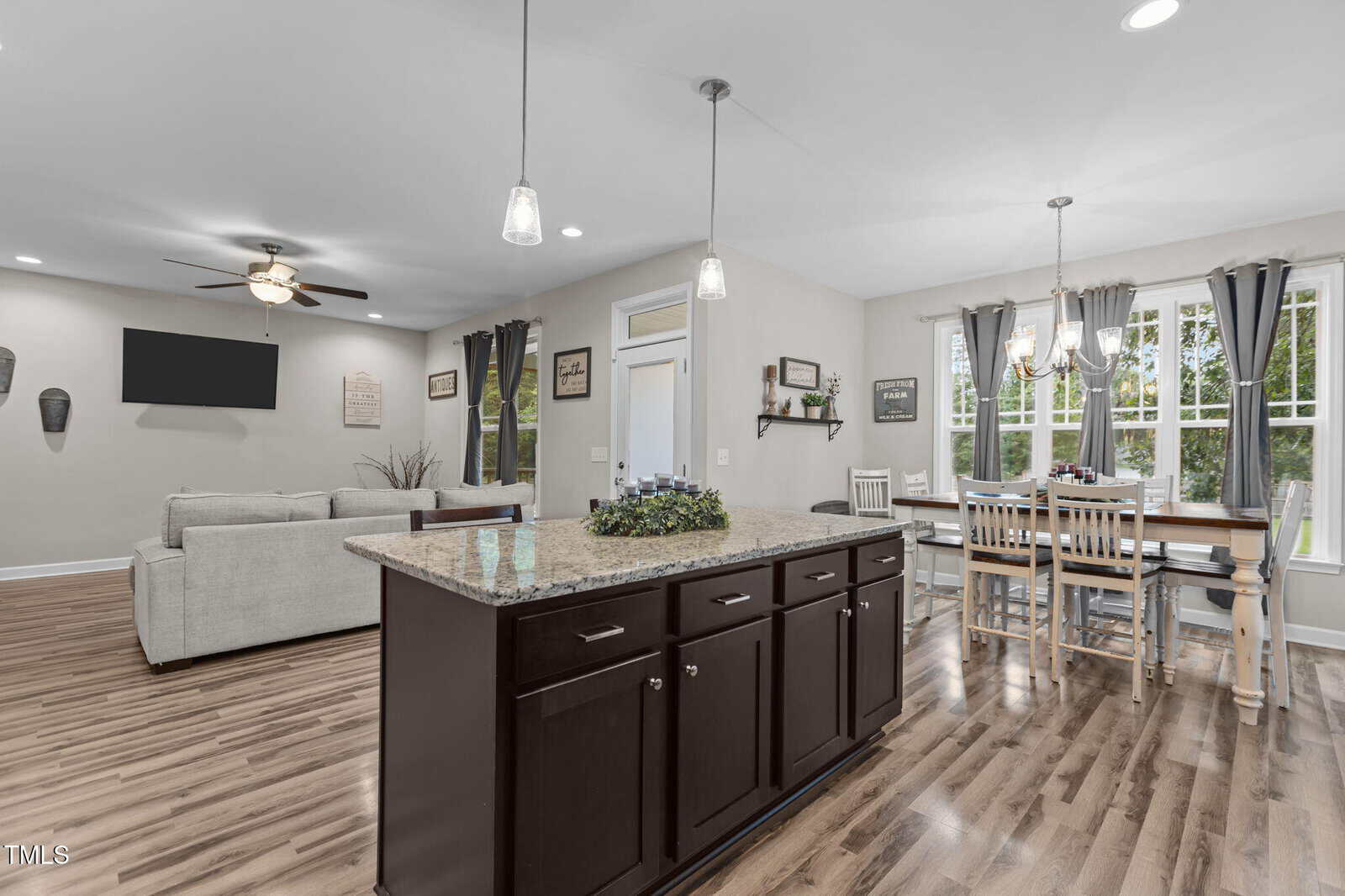 Open-concept kitchen with a granite island, dark cabinets, and pendant lights. Dining area with a wooden table and large windows. Living area with sofas, TV, and ceiling fan. Light walls and wood floors throughout.