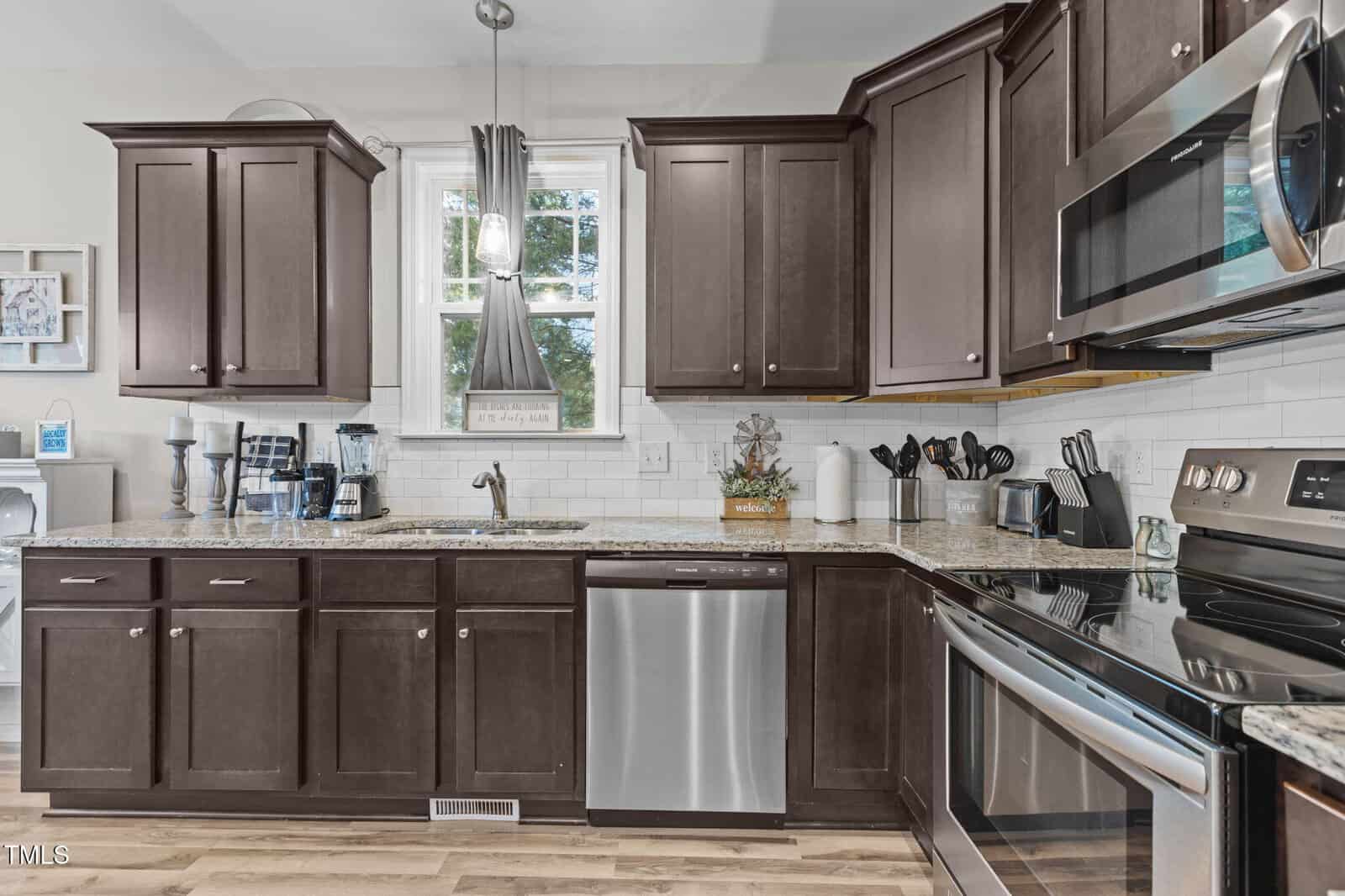 Modern kitchen with dark wood cabinets, stainless steel appliances, granite countertops, a double sink under a window, and white subway tile backsplash. Kitchen utensils and small appliances are neatly arranged on the counters.