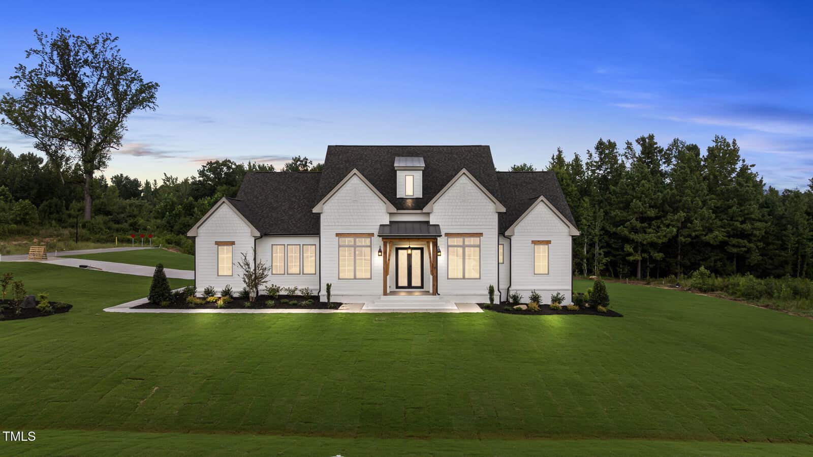 A modern two-story white house with large windows, a dark roof, and a spacious, neatly landscaped green lawn, set against a backdrop of trees under a clear evening sky.
