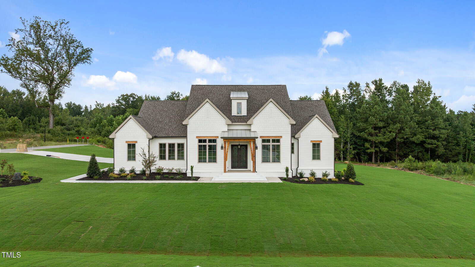 A modern white house with a gabled roof sits on a large, well-manicured green lawn, surrounded by trees under a blue sky with scattered clouds.