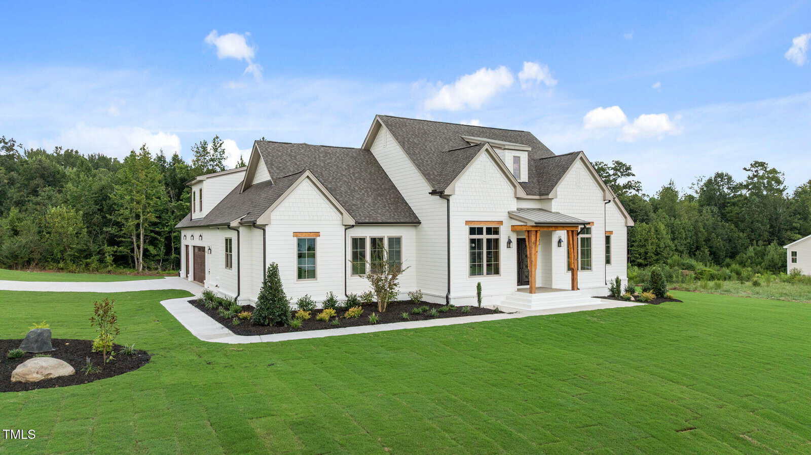 A modern two-story white house with gray roofing, wooden accents, and a covered front porch sits on a large, freshly landscaped lawn with trees and a long driveway. The sky is clear and blue.