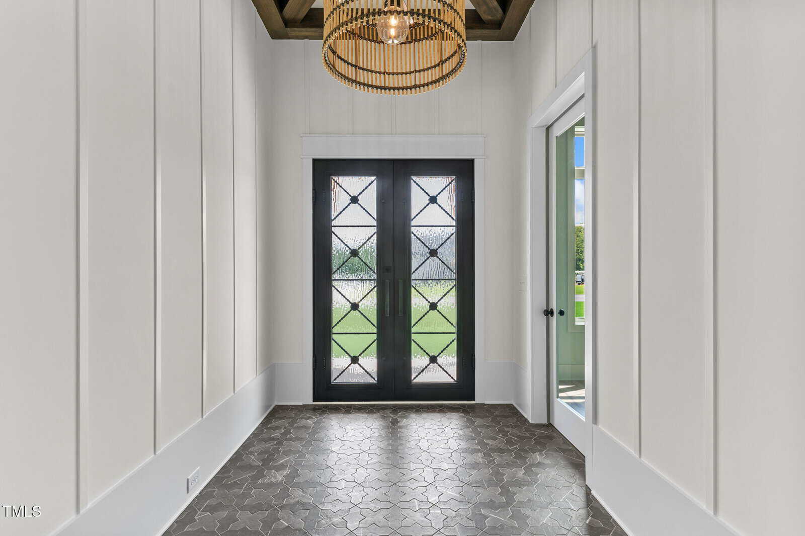 Bright entryway with white vertical wall panels, geometric gray tile floor, black-framed double doors with patterned glass, and a modern chandelier with a wooden ring design hanging from a ceiling with exposed beams.