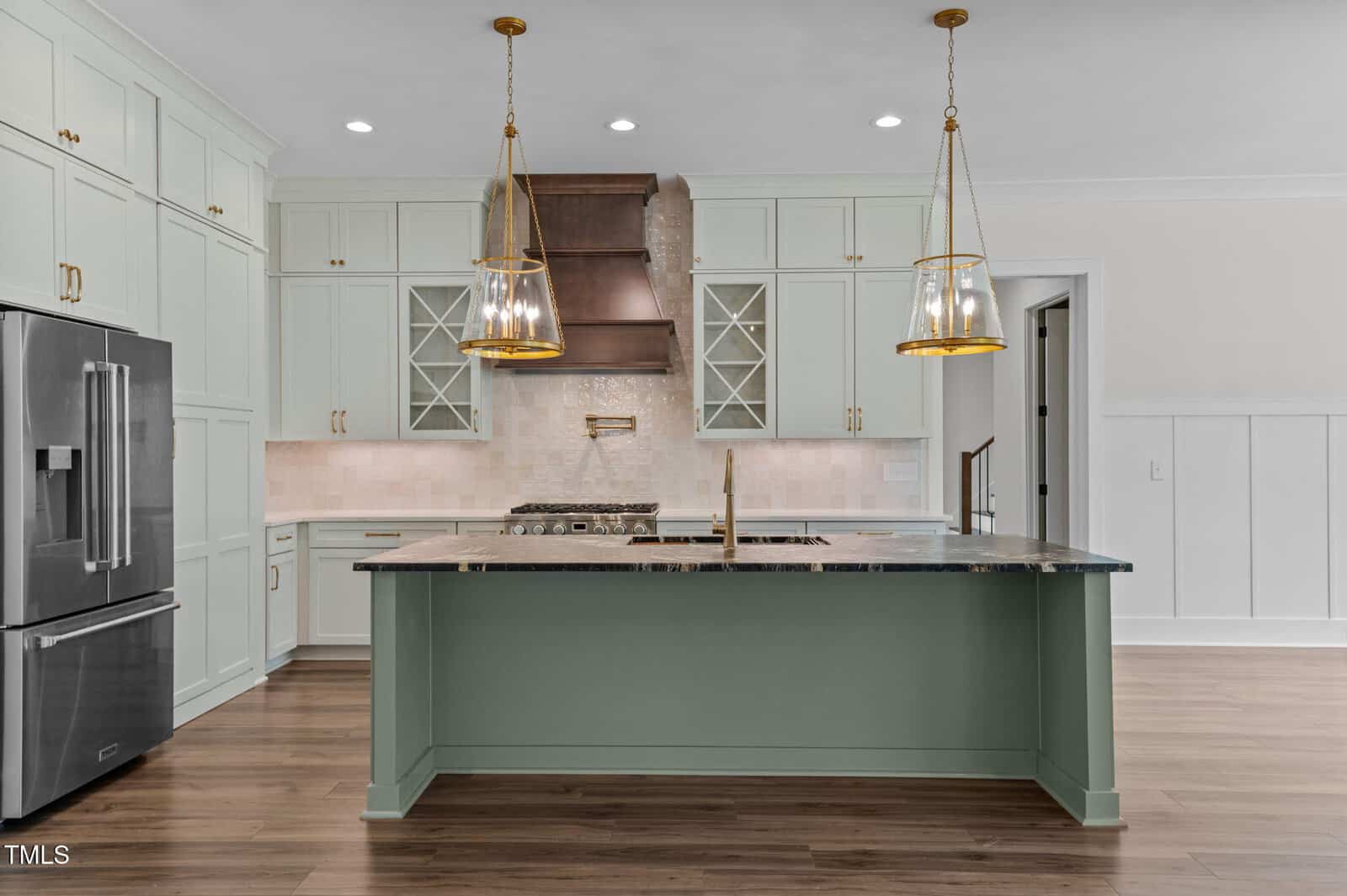 Modern kitchen with light green cabinets, a large island with a dark countertop, stainless steel appliances, two glass pendant lights, and a wood range hood. The floor is wood, and the backsplash is light tile.