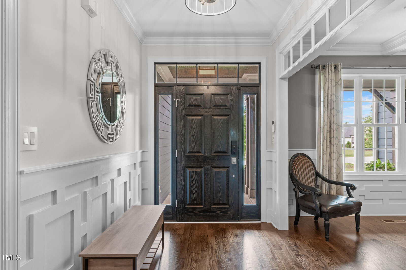 A bright entryway with a dark wooden front door, sidelights, a round decorative mirror on a white paneled wall, a wooden bench, and a classic wooden chair on hardwood floors. Sunlight streams through the glass.