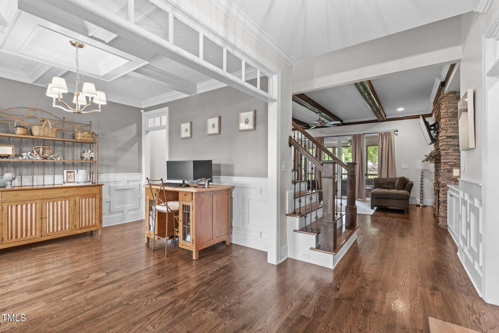A bright, open-concept home interior with hardwood floors, a wooden desk setup, shelving, wainscoting, a wooden staircase with stone accents, and large windows with light curtains in the background.