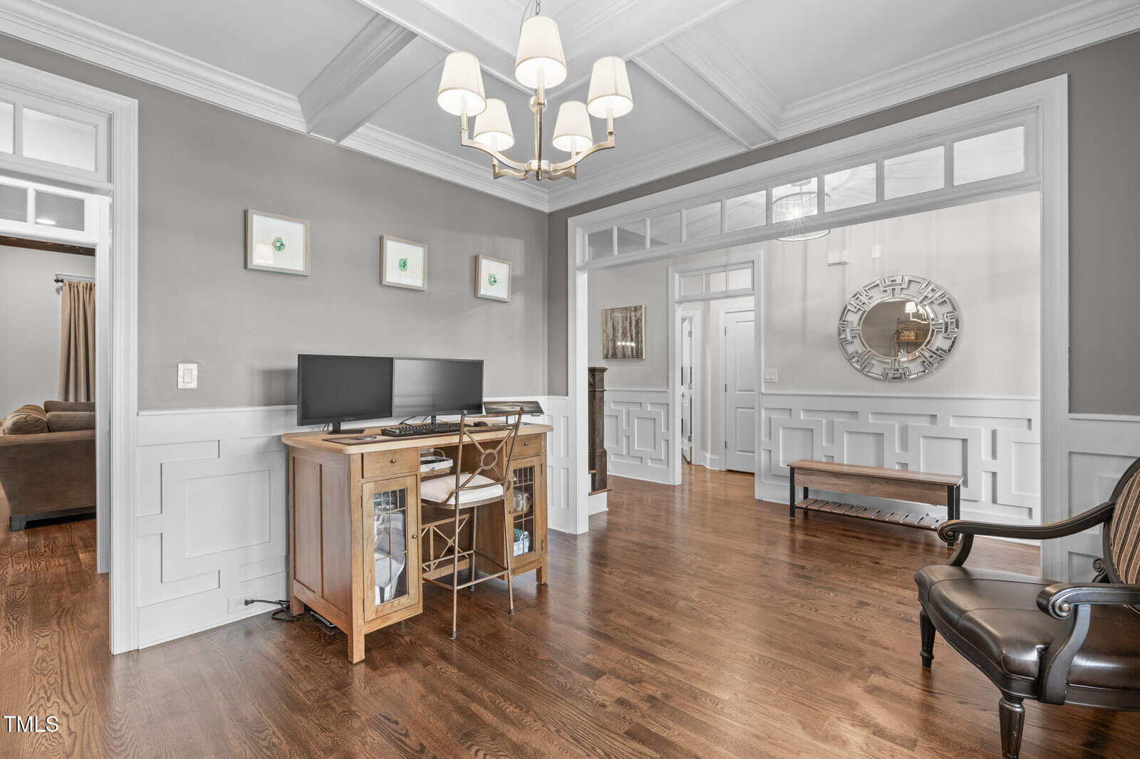 Home office space with dual monitors on a wooden desk, decorative molding, wooden floors, neutral wall color, chandelier above, and a hallway with a bench, mirror, and visible adjacent rooms in the background.