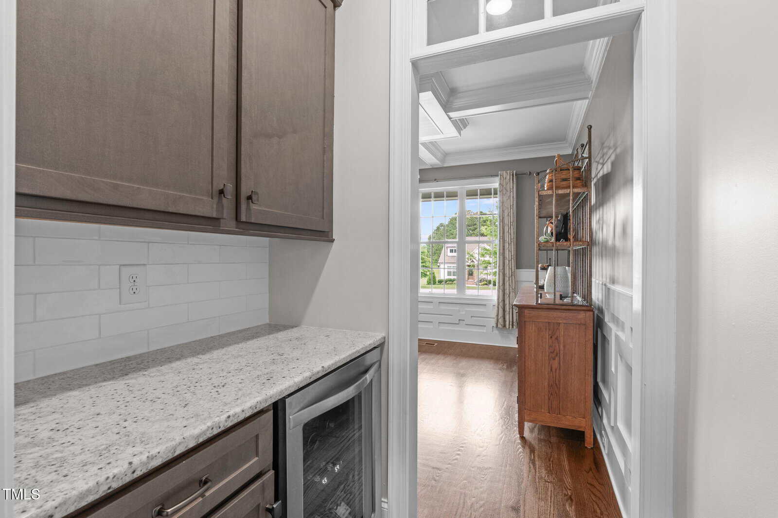 A view from a small kitchen nook with granite countertop and upper cabinets, looking into a bright room with hardwood floors, large windows, and a wooden sideboard.