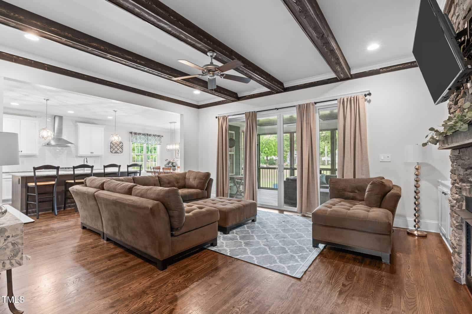 Open living room with brown sectional sofas, a stone fireplace, wooden beam ceiling, and large windows with tan curtains. Hardwood floors and a patterned rug are visible. The space connects to a bright kitchen and dining area.