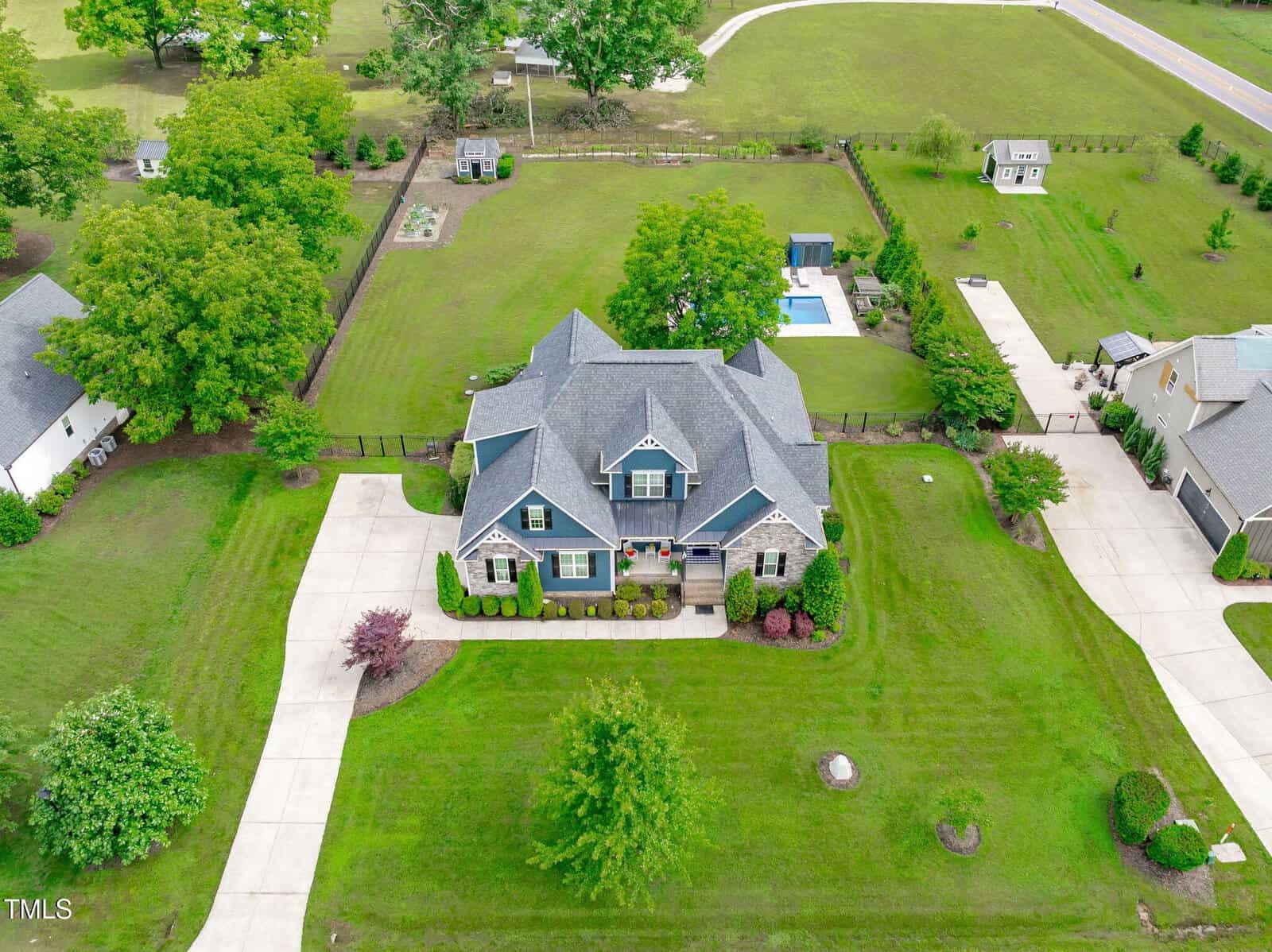 Aerial view of a large gray house with a manicured lawn, trees, and winding driveways. The backyard features a pool, garden, and is bordered by a fence. Neighboring houses and open green space are visible.
