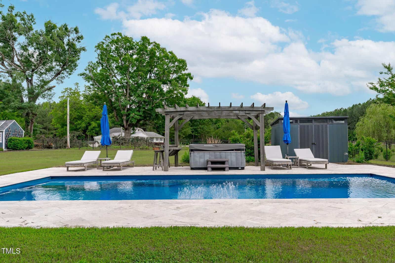 A backyard with a rectangular swimming pool, four lounge chairs with blue umbrellas, a wooden pergola, and a hot tub. Green grass, large trees, and a shed are in the background under a partly cloudy sky.