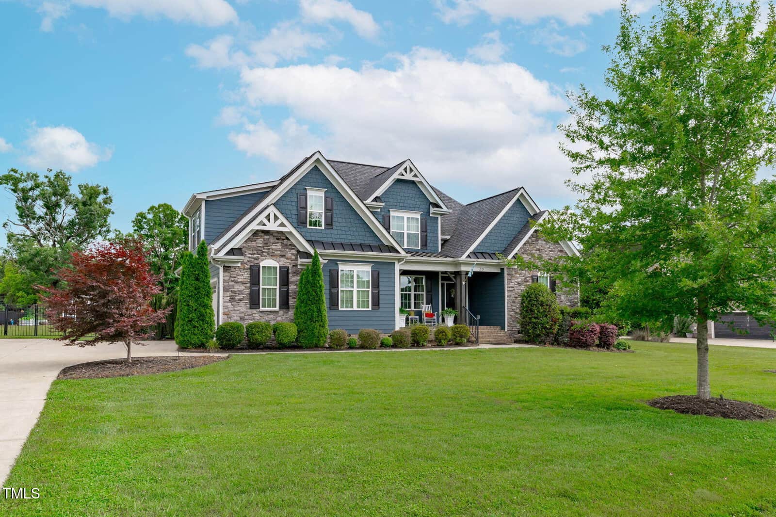 A large modern house with blue siding, stone accents, and multiple gables, surrounded by a well-manicured lawn, trees, and shrubs under a partly cloudy sky.