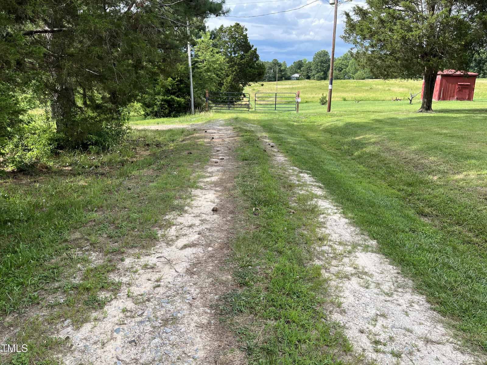A dirt and gravel driveway bordered by grass leads toward a closed metal gate and a green field, with trees on the left and right, and a small red shed visible in the distance.
