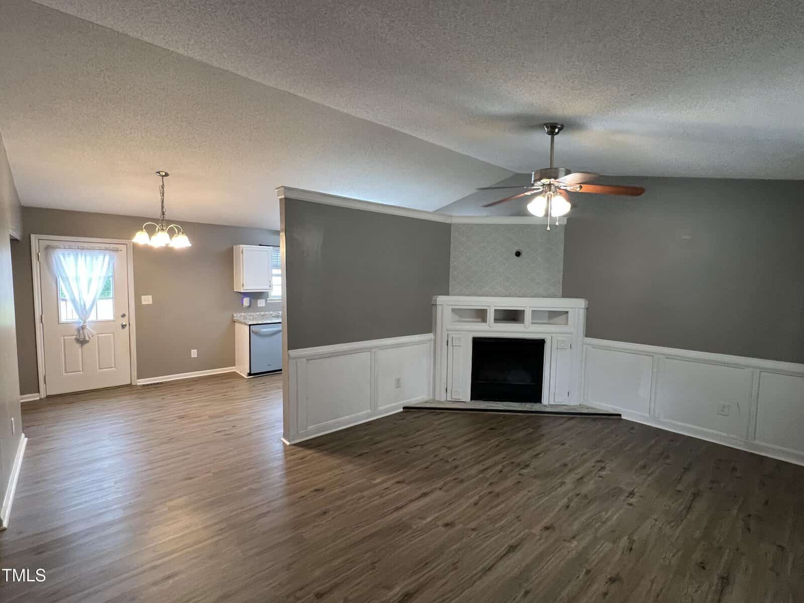 Open living area with wood flooring, gray and white walls, a ceiling fan, a corner fireplace with a white mantel, and an adjacent kitchen and dining area with hanging lights. Natural light enters through a door and window.