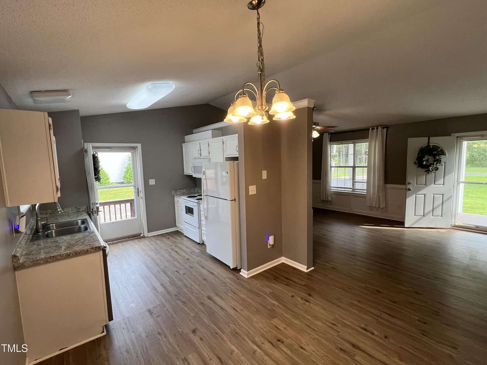 Open kitchen and living area with wood flooring, white cabinets and appliances, a hanging light fixture, and windows allowing natural light. A door leads outside, and the living room has curtains and a wreath on the door.