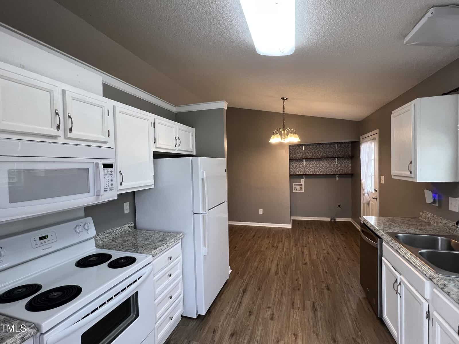 A kitchen with white cabinets, appliances, and countertops, leading to a dining area with a hanging light fixture, wood flooring, and a laundry nook in the back corner.