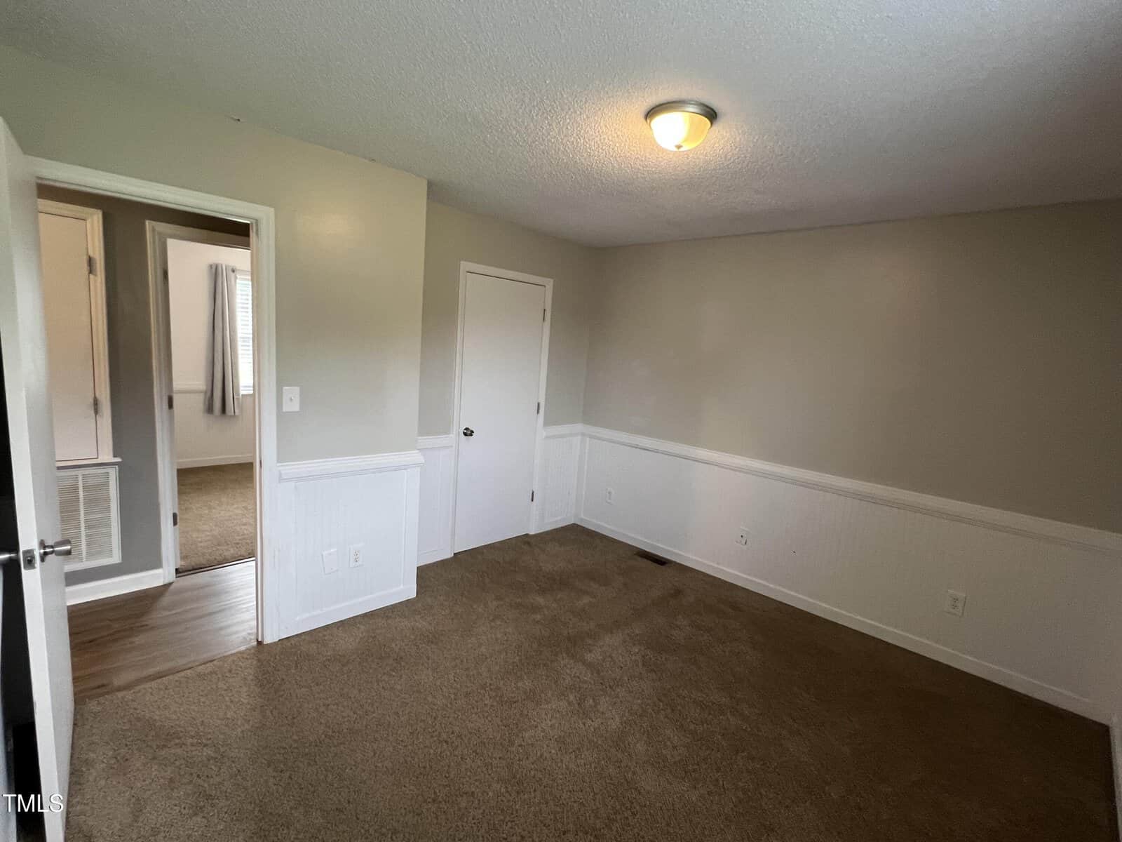 A small, empty room with beige carpet, light-colored walls, a ceiling light, a closed white door, and an open door leading to another room with wood flooring and a window with curtains.