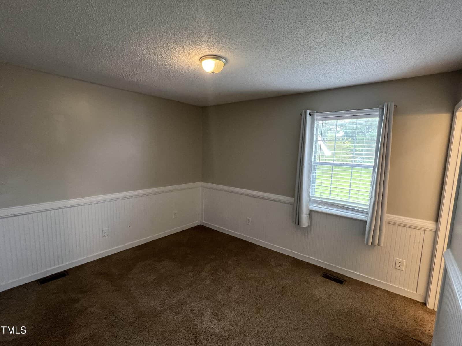 Empty room with beige walls, white wainscoting, brown carpet, a single window with white curtains, and a ceiling light. Natural light enters through the window, revealing a view of green grass outside.
