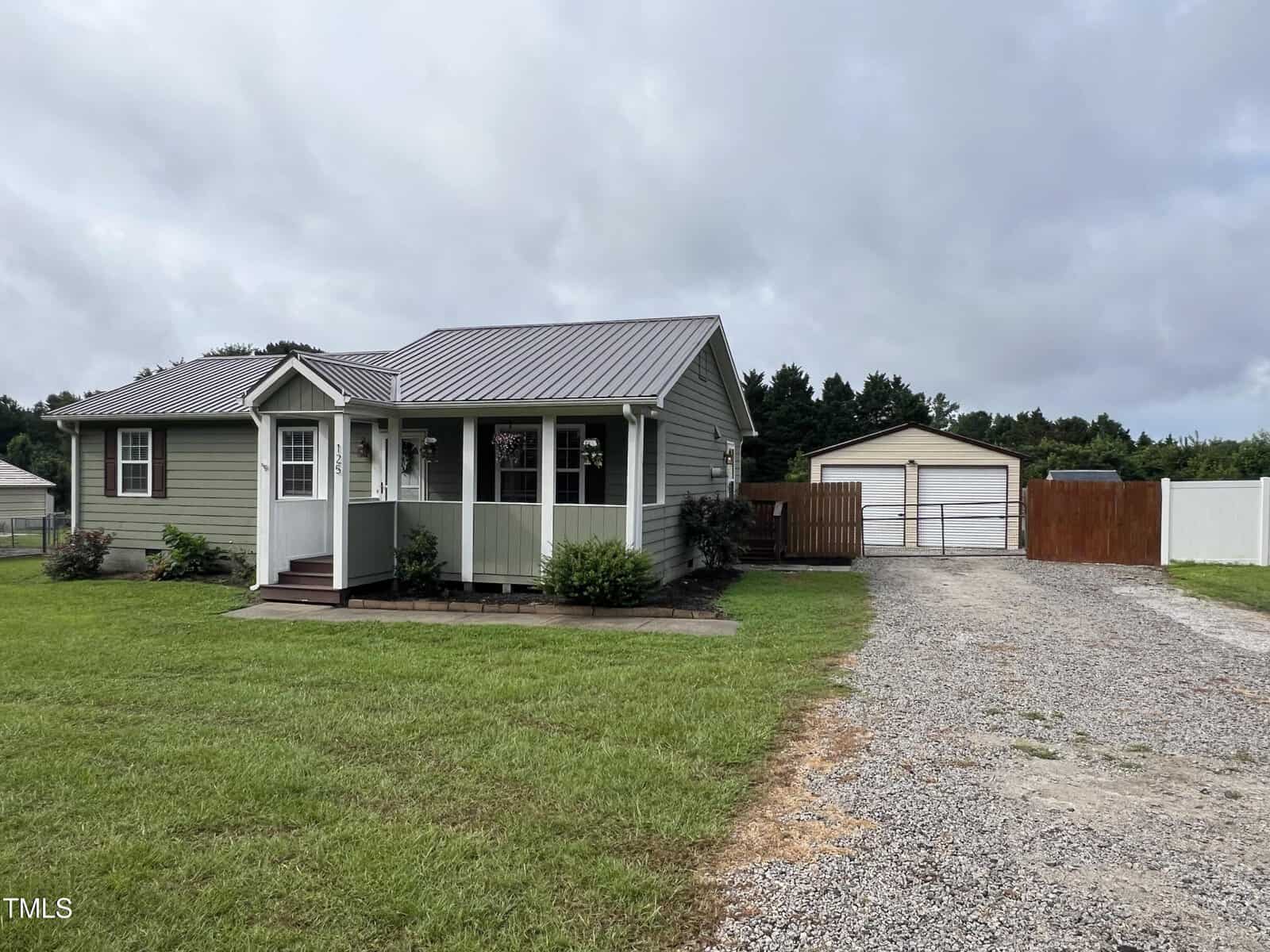 Single-story green house with a small front porch, surrounded by grass. A gravel driveway leads to a detached garage in the background, with trees and a cloudy sky overhead.