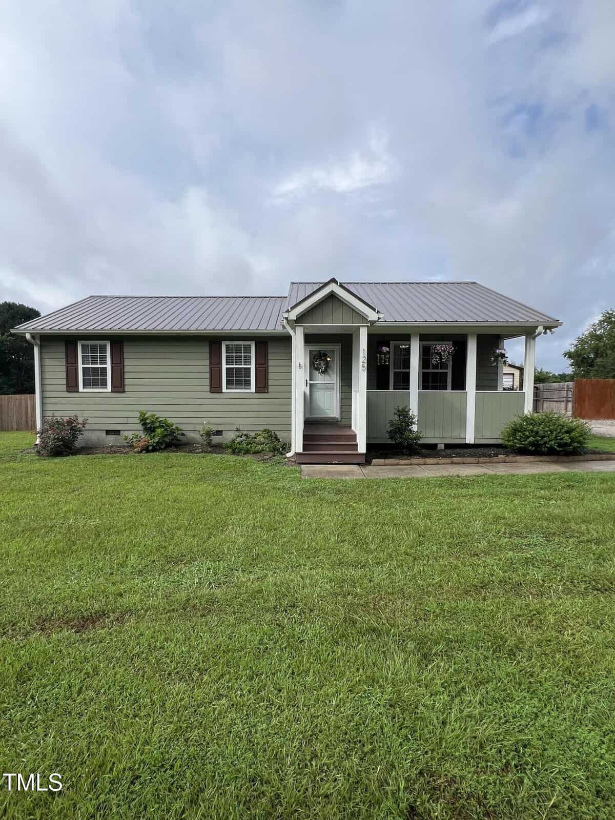 A small, single-story green house with brown shutters and a metal roof, featuring a covered front porch and surrounded by a well-kept lawn and some shrubs.