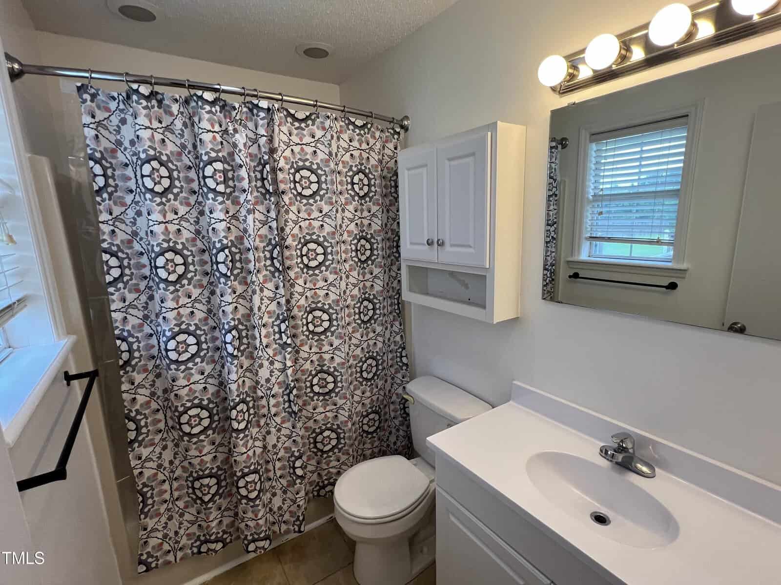 Small bathroom with a patterned shower curtain, white toilet, white vanity with sink, mirrored medicine cabinet, and overhead lighting. Window with blinds above the toilet. Beige tile floor.