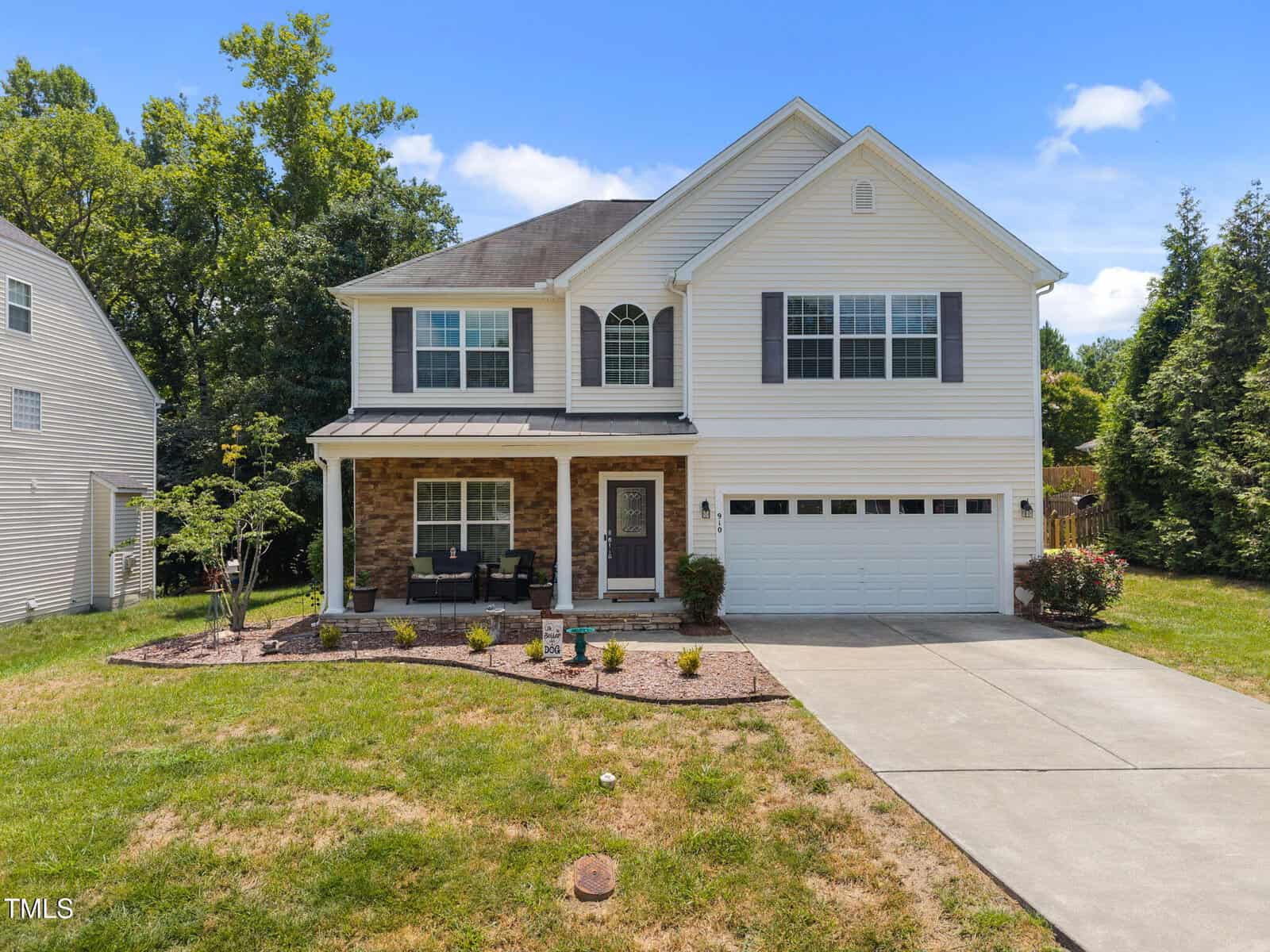 Charming two-story suburban house at 910 Woodland Road in Creedmoor with white siding, dark shutters, a covered front porch with seating, attached two-car garage, concrete driveway, grassy front yard, landscaped garden bed, and small bushes.