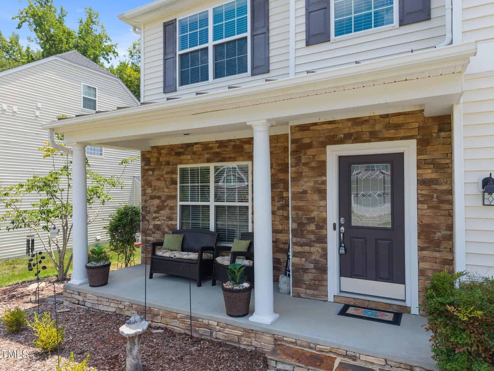 A house with a stone and siding exterior features a covered front porch with two white columns, a black door with a glass panel, a doormat, a bench with cushions, potted plants, and landscaped garden beds.