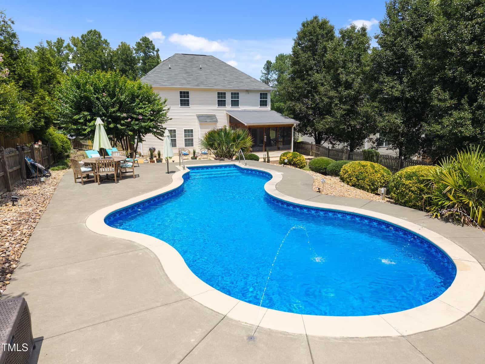 A backyard with a curvy in-ground swimming pool, water jets, a concrete patio, patio furniture with an umbrella, landscaped bushes, and a two-story house in the background under a blue sky.